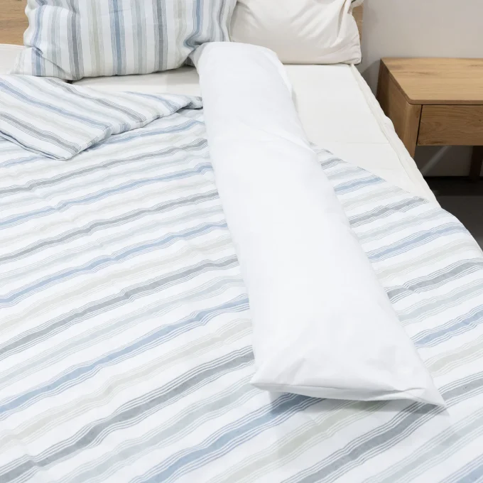 Housse anti-acariens traversin Softsan Protect Plus posée sur un lit avec linge rayé